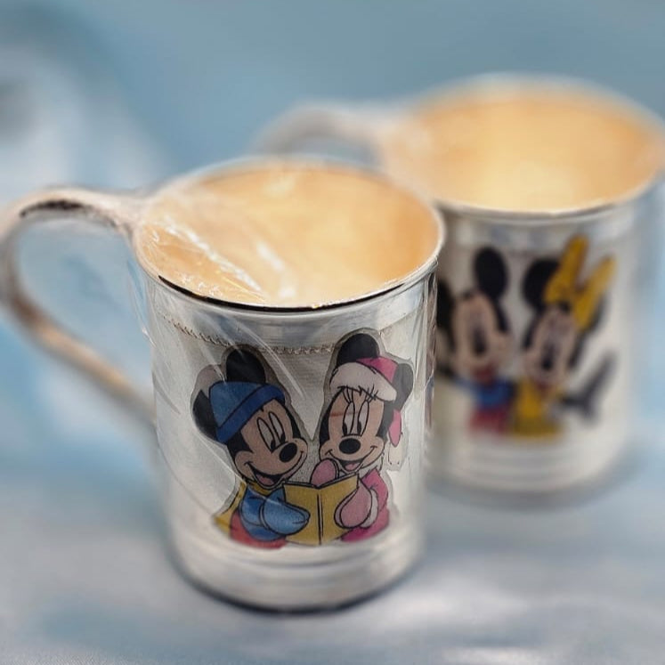 Two silver mugs with cartoon character designs on a light blue fabric background