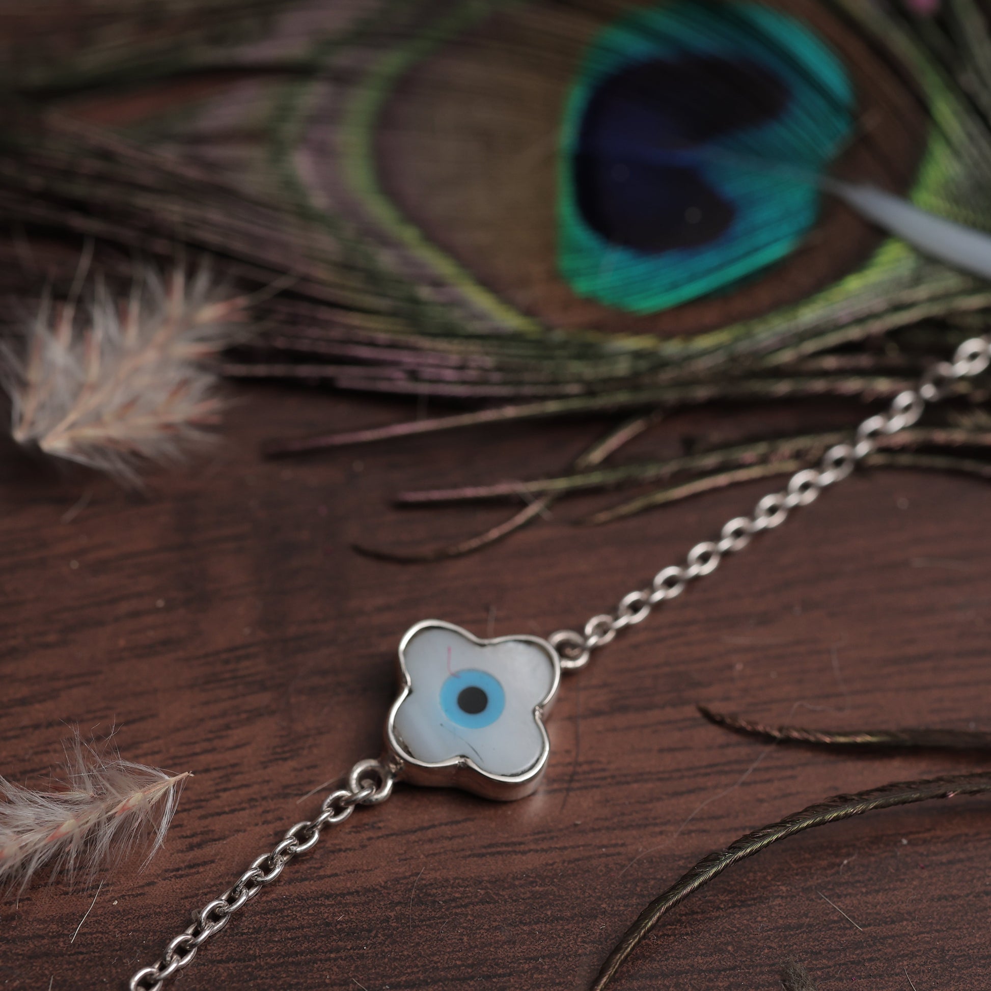 Silver bracelet with a clover-shaped charm on a wooden surface with peacock feathers.