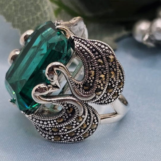 Decorative ring with green gemstone and intricate silver design on a light blue fabric background